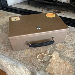 May include: A brown metal box with a black handle and a key. The box has a lock and two stickers on the top. The key is attached to a white string. The box is sitting on a stone surface.