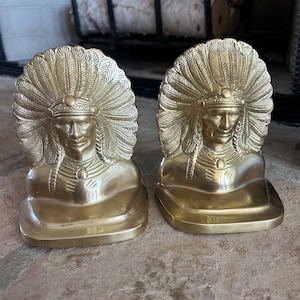 May include: Two brass bookends, each featuring a detailed bust of a Native American chief wearing a feathered headdress. The bookends have a gold finish and are placed on a stone surface.