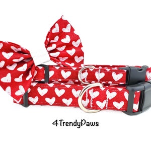 May include: Red dog collar and bow tie with a white heart pattern. The collar has a black buckle and a silver D-ring. The bow tie is made of fabric and has a white bow. The text "4 Trendy Paws" is visible at the bottom of the image.
