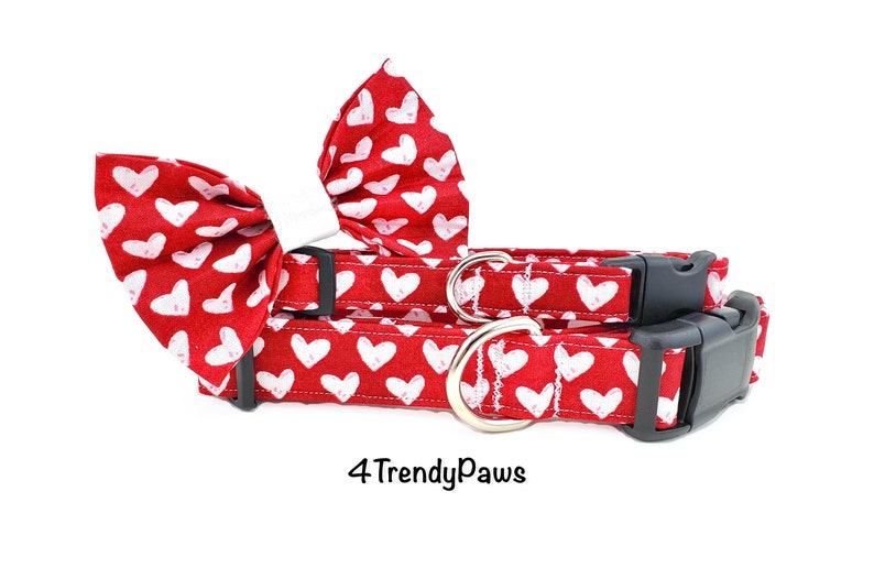May include: Red dog collar and bow tie with a white heart pattern. The collar has a black buckle and a silver D-ring. The bow tie is made of fabric and has a white bow. The text "4 Trendy Paws" is visible at the bottom of the image.