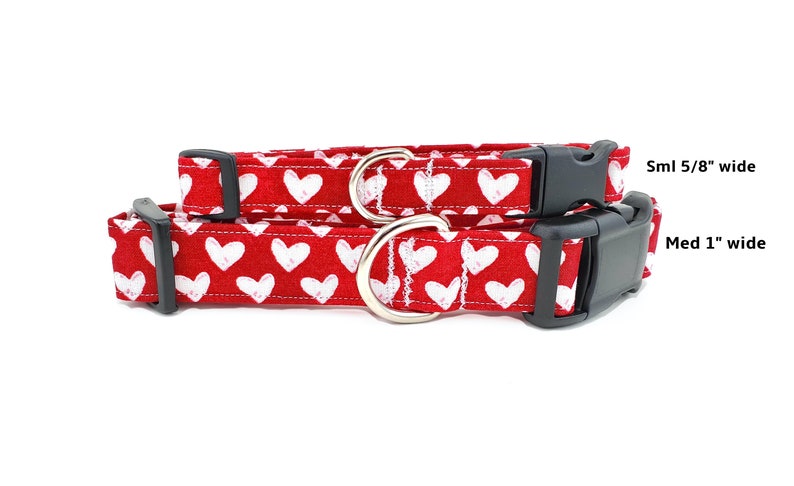 May include: Two red dog collars with white hearts printed on them. The smaller collar is 5/8 inch wide and the larger collar is 1 inch wide. Both collars have black plastic buckles.