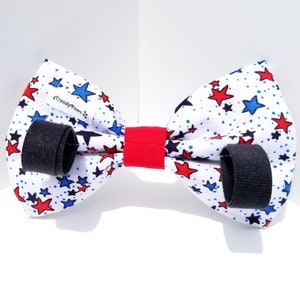 May include: A white bow tie with red and blue stars on a white background. The bow tie has black adjustable straps and a red center piece. The bow tie has the text "Trendy Paws" on it.