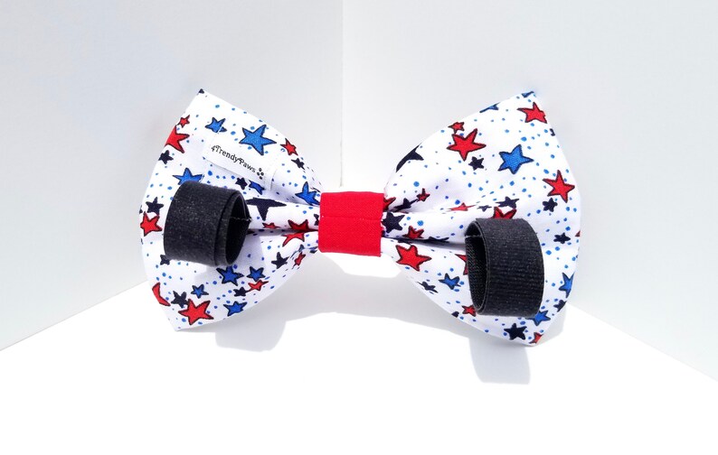 May include: A white bow tie with red and blue stars on a white background. The bow tie has black adjustable straps and a red center piece. The bow tie has the text "Trendy Paws" on it.