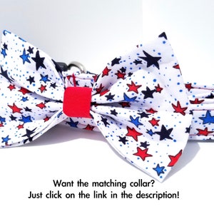 May include: A white dog collar with a red, white, and blue star print bow tie. The bow tie is made of fabric and has a red center. The collar is adjustable and has a black buckle.