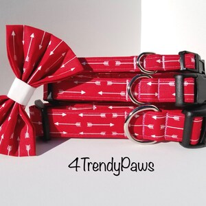 May include: Three red dog collars with a white arrow print and black buckles. The collars are stacked on top of each other. A matching red bow tie with a white arrow print is in the foreground. The text "4 Trendy Paws" is visible below the collars.