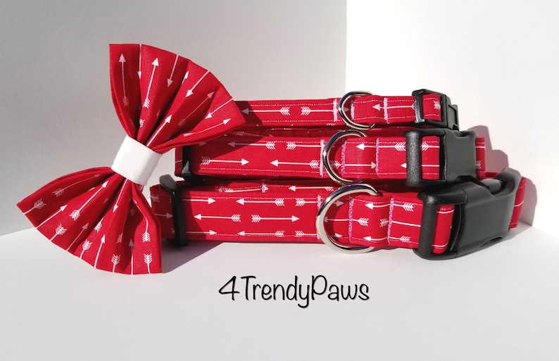 May include: Three red dog collars with a white arrow print and black buckles. The collars are stacked on top of each other. A matching red bow tie with a white arrow print is in the foreground. The text "4 Trendy Paws" is visible below the collars.