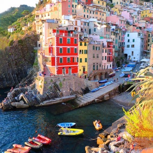 May include: A colourful village built on a cliffside overlooking a harbour with boats in the water. The buildings are painted in shades of red, yellow, orange, pink, and white.