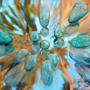 May include: A collection of teal blue glass stones arranged in a circular pattern on a reflective surface. The stones are smooth and have a slight shimmer to them. The background is a blurred image of orange and blue colors.