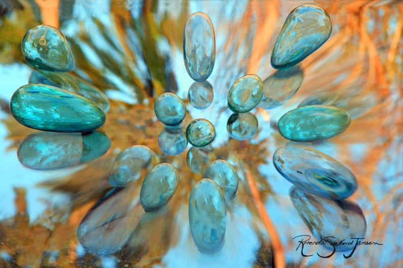 May include: A collection of teal blue glass stones arranged in a circular pattern on a reflective surface. The stones are smooth and have a slight shimmer to them. The background is a blurred image of orange and blue colors.
