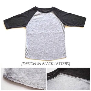 May include: A gray and black baseball tee shirt with a raglan sleeve design. The shirt has a black crew neck and black sleeves. The text "DESIGN IN BLACK LETTERS" is printed on the bottom of the shirt.