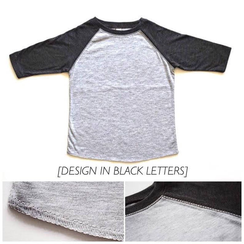 May include: A gray and black baseball tee shirt with a raglan sleeve design. The shirt has a black crew neck and black sleeves. The text "DESIGN IN BLACK LETTERS" is printed on the bottom of the shirt.
