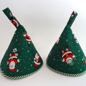 May include: Two green Christmas pot holders with a festive Santa Claus print. The pot holders have a cone shape with a loop for hanging and a striped border. The Santa Claus is depicted playing sports like soccer and hockey.