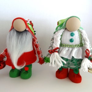 May include: Two handmade Christmas gnomes. One gnome wears a red and white checkered hat and coat, a long white beard, and green shoes. The other gnome has a green and white hat, white braided hair, and a white and green dress. Both gnomes hold bells.