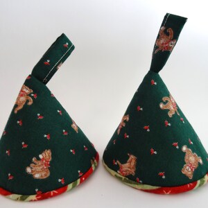 May include: Two cone-shaped fabric items with a dark green background, adorned with teddy bear and holly berry patterns. Each has a red and green patterned trim and a fabric loop at the top. The items are likely decorative or functional.