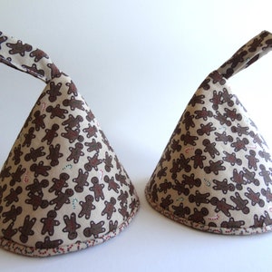 May include: Two cone-shaped fabric tea cozies. The fabric features a repeating pattern of brown gingerbread men and candy canes on a beige background. Each cozy has a matching fabric handle. The edges are trimmed with a red, white, and green dotted border.