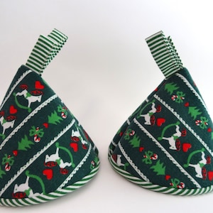 May include: Two green Christmas tree-shaped pot holders with a festive pattern of rocking horses, hearts, and trees. Each pot holder has a striped green and white ribbon loop for hanging and a striped border.