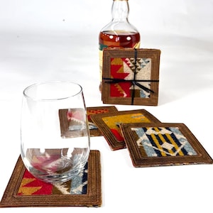 May include: Set of six colourful coasters with a geometric pattern. The coasters are made of a woven fabric with a brown leather border. A glass and a bottle of spirits are on the table.