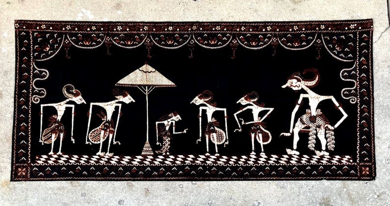 Op de afbeelding: Een zwart-witte wandhanger van textiel met een traditioneel Indonesisch ontwerp. De hanger toont een sc&egrave;ne met meerdere figuren, een parasol en ingewikkelde patronen.