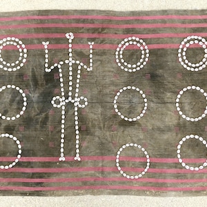 Könnte beinhalten: Ein braunes Stoffgewebe mit einem geometrischen Muster, das mit weißen Kaurischneckenmuscheln geschaffen wurde. Das Muster zeigt Kreise und Linien. Der Stoff hat einen rosa-weißen gestreiften Rand.