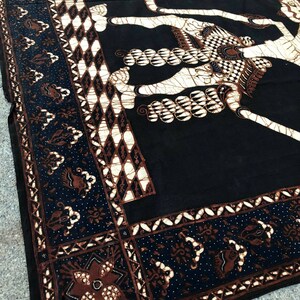Op de afbeelding: Zwarte en bruine batikstof met een wit ontwerp van een mythisch wezen. De stof heeft een decoratieve rand met een herhalend patroon.
