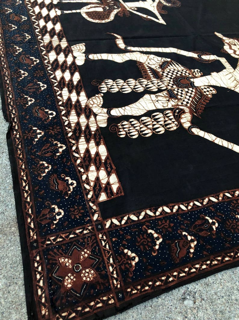 Op de afbeelding: Zwarte en bruine batikstof met een wit ontwerp van een mythisch wezen. De stof heeft een decoratieve rand met een herhalend patroon.