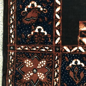 Op de afbeelding: Een zwarte stof met een bruin en wit batikpatroon. Het patroon heeft een bloemmotief met een centrale bloem en een geometrische rand.