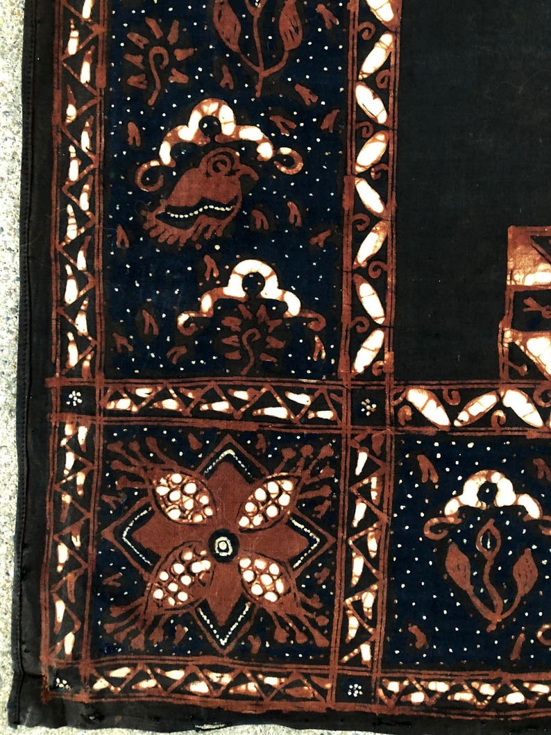 Op de afbeelding: Een zwarte stof met een bruin en wit batikpatroon. Het patroon heeft een bloemmotief met een centrale bloem en een geometrische rand.