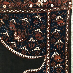 Op de afbeelding: Zwarte stof met een wit en bruin batikpatroon. Het patroon bevat gestileerde vogels, bloemen en geometrische vormen. De stof heeft een textuur.