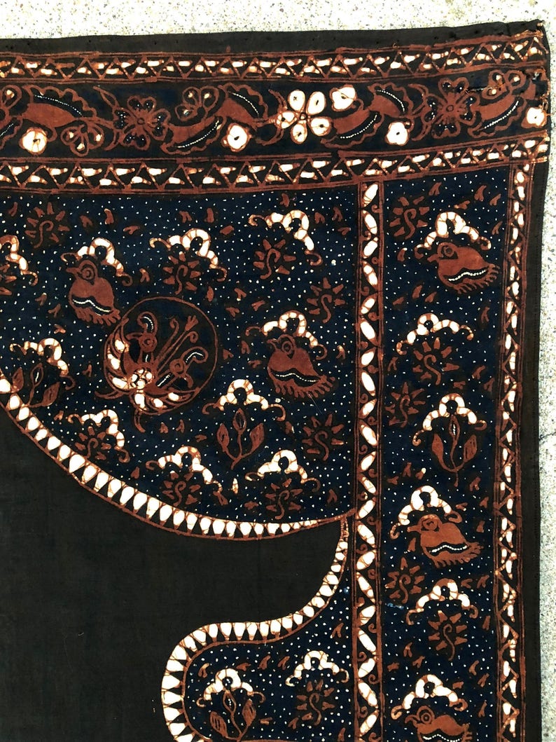 Op de afbeelding: Zwarte stof met een wit en bruin batikpatroon. Het patroon bevat gestileerde vogels, bloemen en geometrische vormen. De stof heeft een textuur.