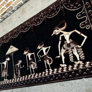 Op de afbeelding: Zwarte en witte textiel wandhanger met een traditioneel Indonesisch ontwerp met figuren in een processie. De figuren zijn gestileerd en hebben een poppenachtig uiterlijk. De achtergrond is zwart met witte details.