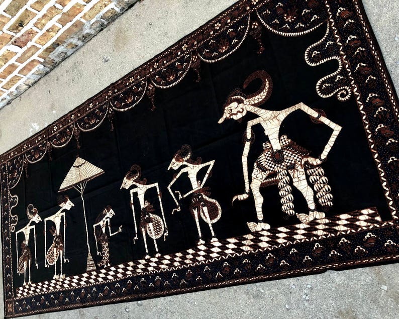 Op de afbeelding: Zwarte en witte textiel wandhanger met een traditioneel Indonesisch ontwerp met figuren in een processie. De figuren zijn gestileerd en hebben een poppenachtig uiterlijk. De achtergrond is zwart met witte details.