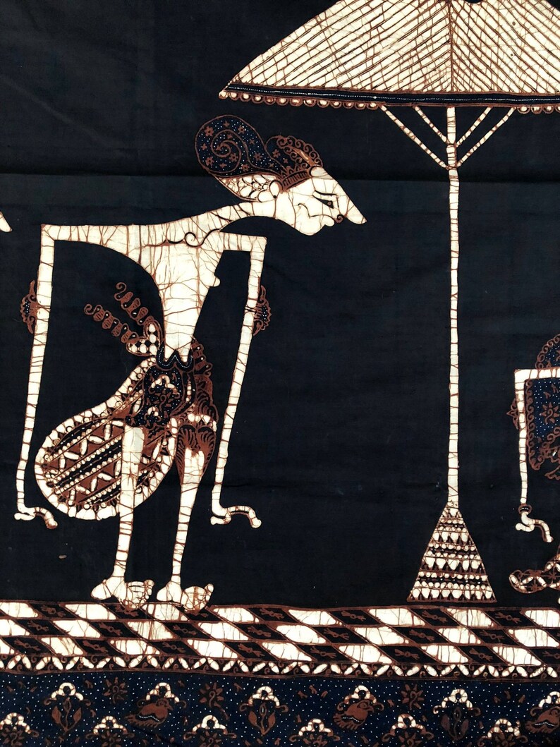 Op de afbeelding: Een zwart-witte batikstof met een traditioneel Indonesisch poppenontwerp. De pop houdt een schild vast en staat onder een witte paraplu. De stof heeft een bruin-witte rand.