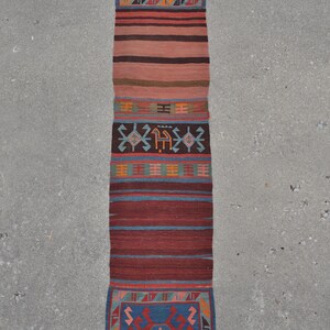 May include: A long, narrow rug with a geometric pattern in shades of red, blue, and orange. The rug has a fringe on both ends.