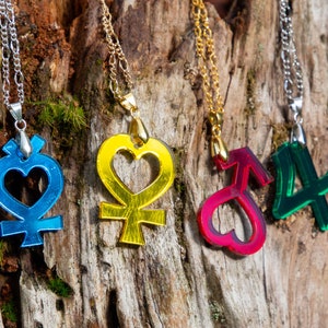 Puede incluir: Cinco collares coloridos con diferentes dijes colgando de una superficie de madera. Los dijes son un corazón azul con un símbolo, un corazón amarillo con un símbolo, un corazón rojo con un símbolo, un número cuatro verde y un símbolo rojo con un corazón.