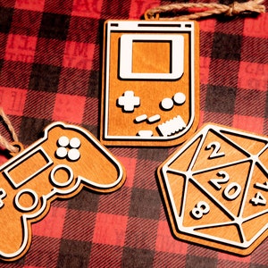May include: Three wooden ornaments shaped like a video game controller, a games console, and a 20-sided die. The ornaments are painted brown with white details.
