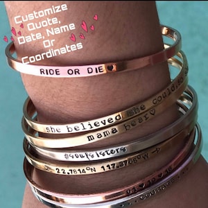 May include: A stack of gold and silver bracelets with various engraved messages, including "Ride or Die", "She believed she could, so mama bear", "#souls sisters", "22.7814°N 117.3768°W", "01 10 16", and "Courage".