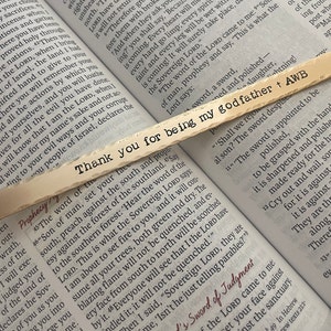 May include: A close-up of a wooden bookmark with the text "Thank you for being my godfather + AWB" written on it. The bookmark is lying on top of an open book with text about swords and judgment.