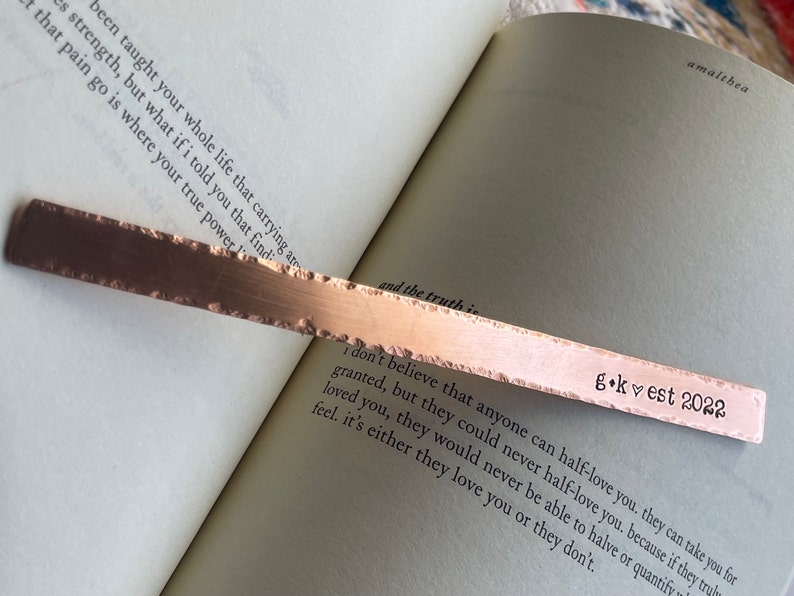May include: A copper bookmark with a rough, hammered texture. The bookmark has the text "g.k. &hearts; est 2022" engraved on it. The bookmark is lying on a book with a quote about love and truth.