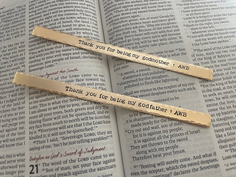 Op de afbeelding: Twee houten bladwijzers met de tekst "Bedankt dat je mijn peter bent" en "Bedankt dat je mijn meter bent". De bladwijzers liggen op een open boek met tekst over de Bijbel.