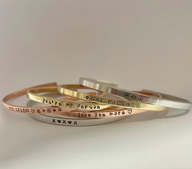 May include: A set of four gold, silver, and copper bracelets with different engraved messages. The messages include "my tribe", "you're my person", "gal pals", and "love you more".