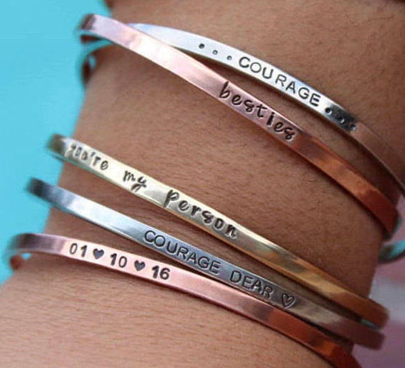 May include: A stack of five silver, gold, and copper bracelets with engraved messages. The bracelets read "Courage", "Besties", "You're my person", "Courage Dear", and "01 10 16".
