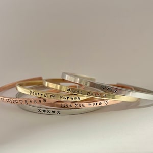 May include: Five stacked gold, silver, and rose gold bracelets with engraved messages. The messages read "My tribe", "You're my person", "Gal pals", "Love you more", and "K+E+A".