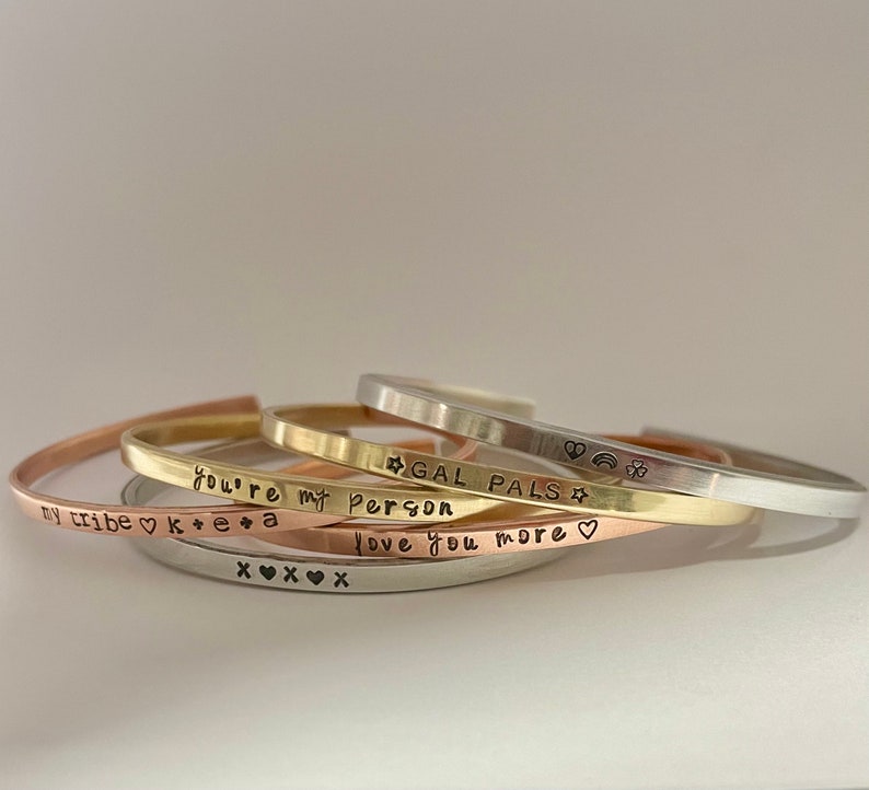 May include: Five stacked gold, silver, and rose gold bracelets with engraved messages. The messages read "My tribe", "You're my person", "Gal pals", "Love you more", and "K+E+A".