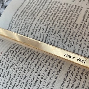 May include: A close-up of a gold bookmark with the text "Amor Fati" printed on it. The bookmark is resting on an open book with text about the Bible.