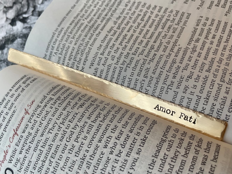 May include: A close-up of a gold bookmark with the text "Amor Fati" printed on it. The bookmark is resting on an open book with text about the Bible.