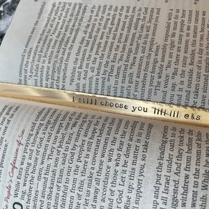 May include: A gold bookmark with the words "i still choose you" engraved on it. The bookmark is decorated with a series of vertical lines.