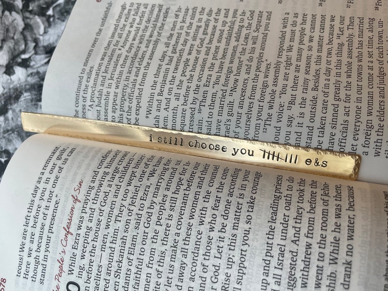 May include: A gold bookmark with the words "i still choose you" engraved on it. The bookmark is decorated with a series of vertical lines.