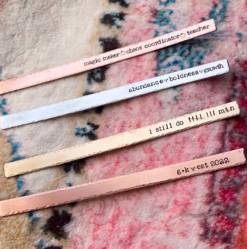 May include: Four metal bars with different colored finishes and engraved text. The text on the bars reads: "magic maker chaos coordinator teacher", "abundance boldness growth", "i still do HL 111 m&n", and "g+kvest 2022".