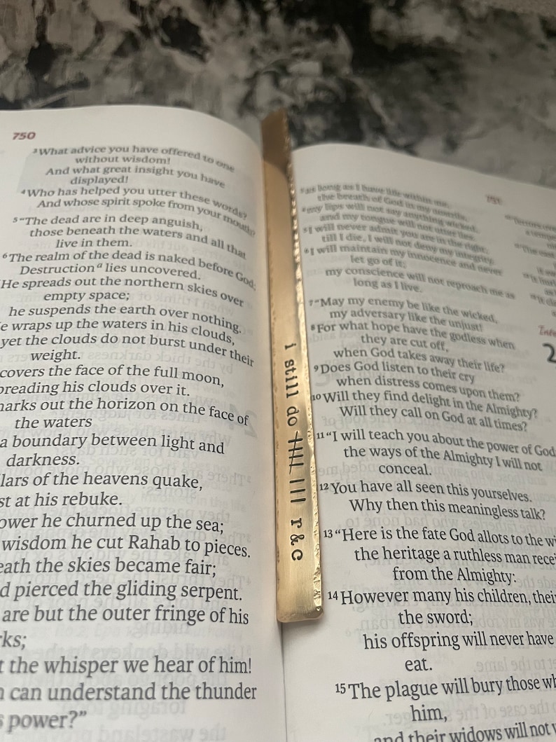 May include: A gold bookmark with the words "I still do" and "R & C" engraved on it. The bookmark is placed on a page of a book with text about the book of Job in the Bible.