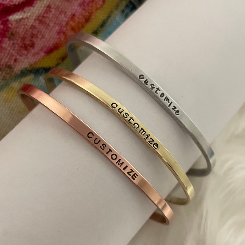 May include: Three minimalist bangle bracelets in silver, gold, and rose gold with the word "CUSTOMIZE" engraved on each.